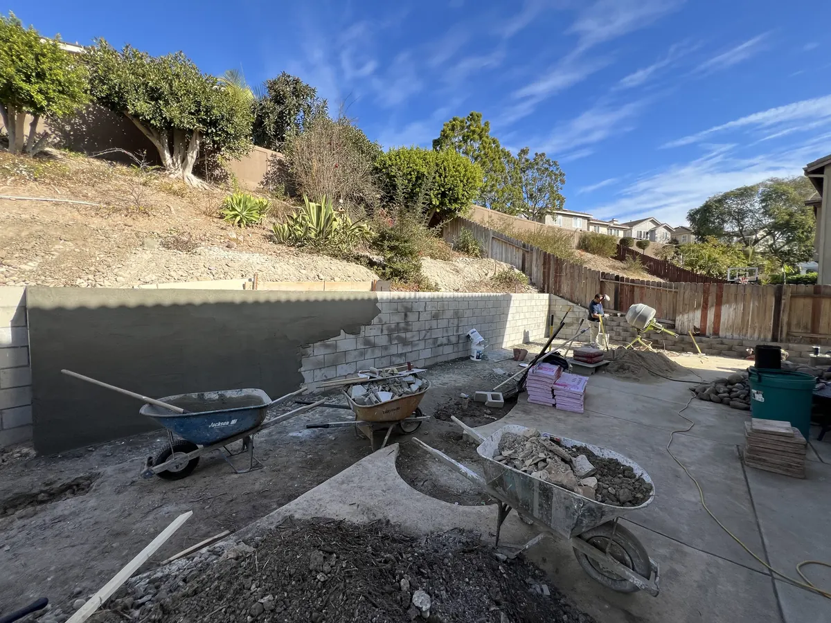Concrete block retaining wall going up across the back of the yard, partly finished with a smooth coat
