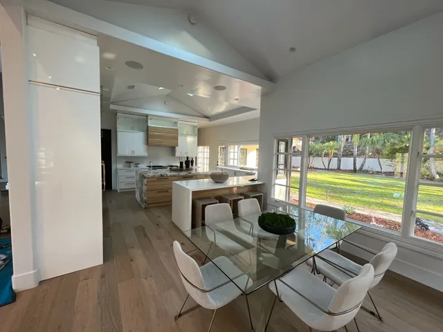 Bright open kitchen and dining area with wood floors, white island, marble countertops, and a large picture window showing a green lawn.