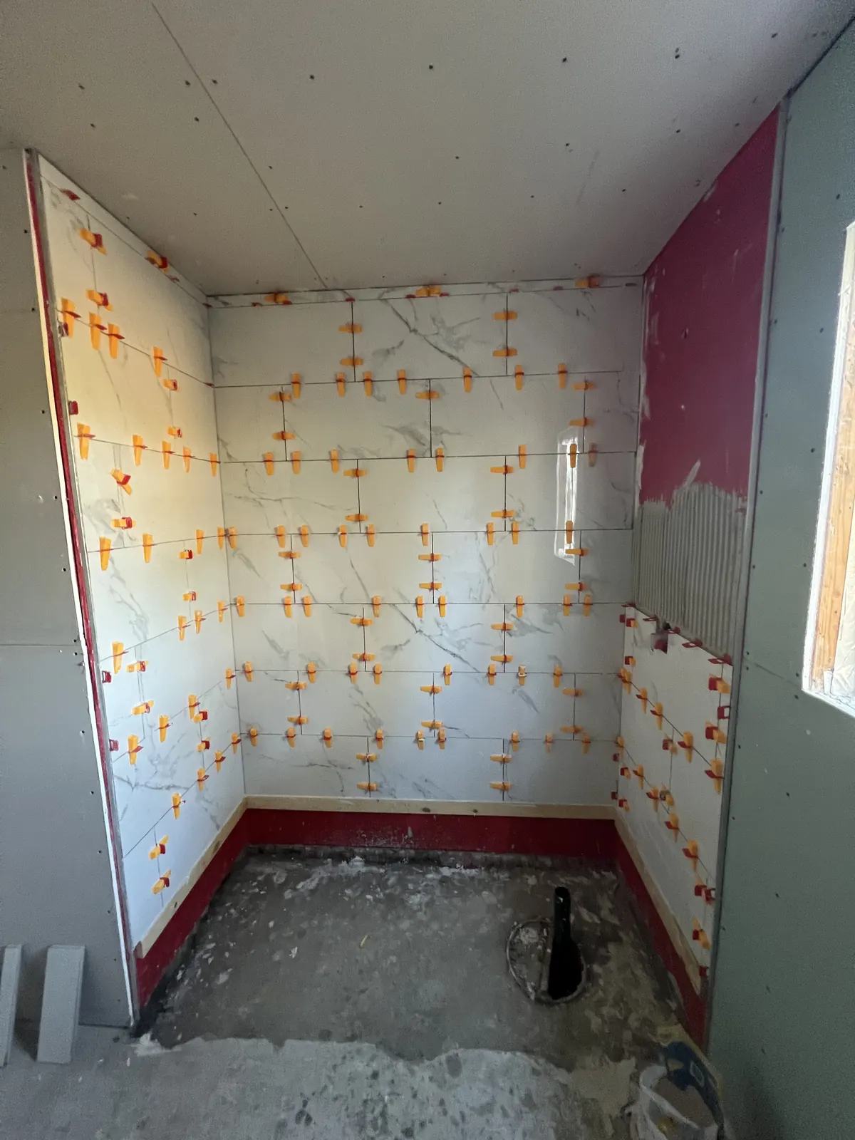 Large marble-look tile being set with orange leveling clips over a red liquid waterproofing coat