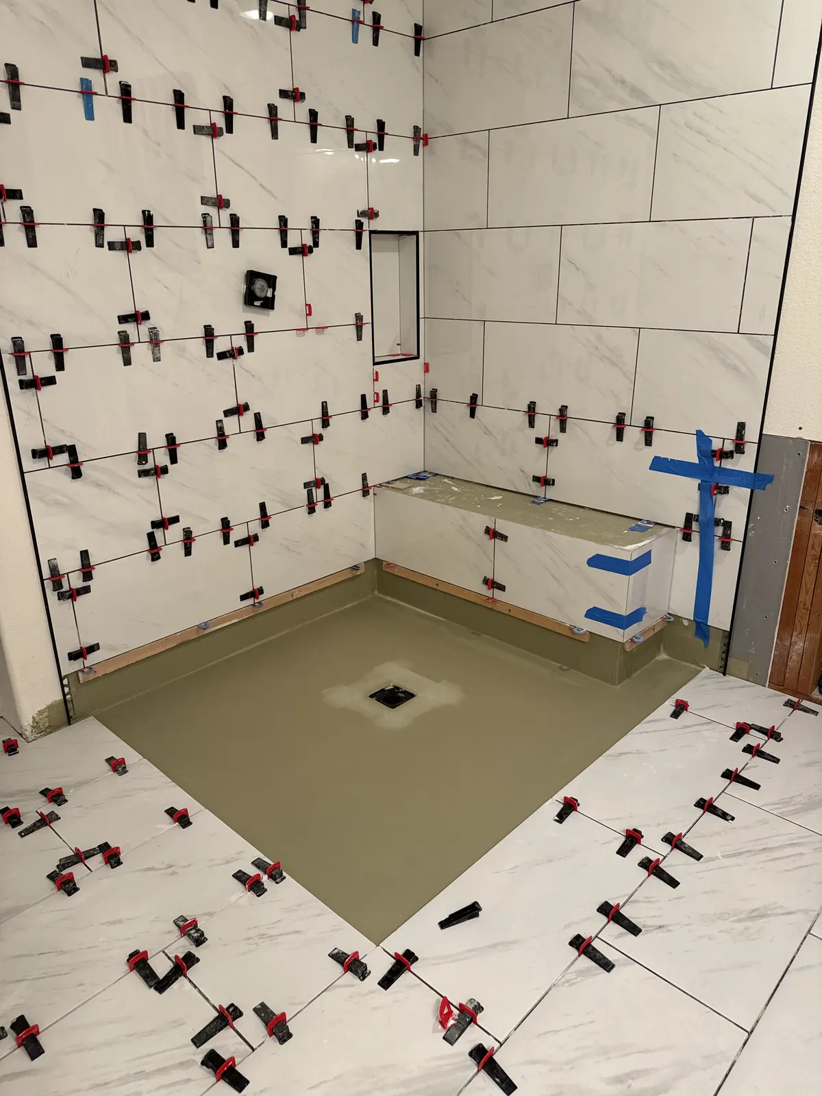 Large marble-look tiles being set with leveling clips around a sloped shower pan and bench