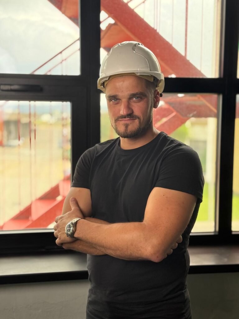 Confident construction professional wearing a white hard hat stands with arms crossed in front of a large window on a job site. About Us — Construction Crafters team member on site
