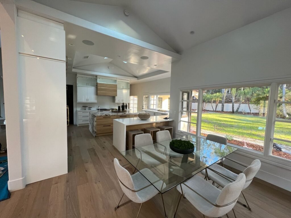 Bright open kitchen and dining area with wood floors, glass dining table with six chairs, white island with bar stools, marble countertops, upper cabinets, and a large picture window showing a green lawn outside — Construction Crafters project.
