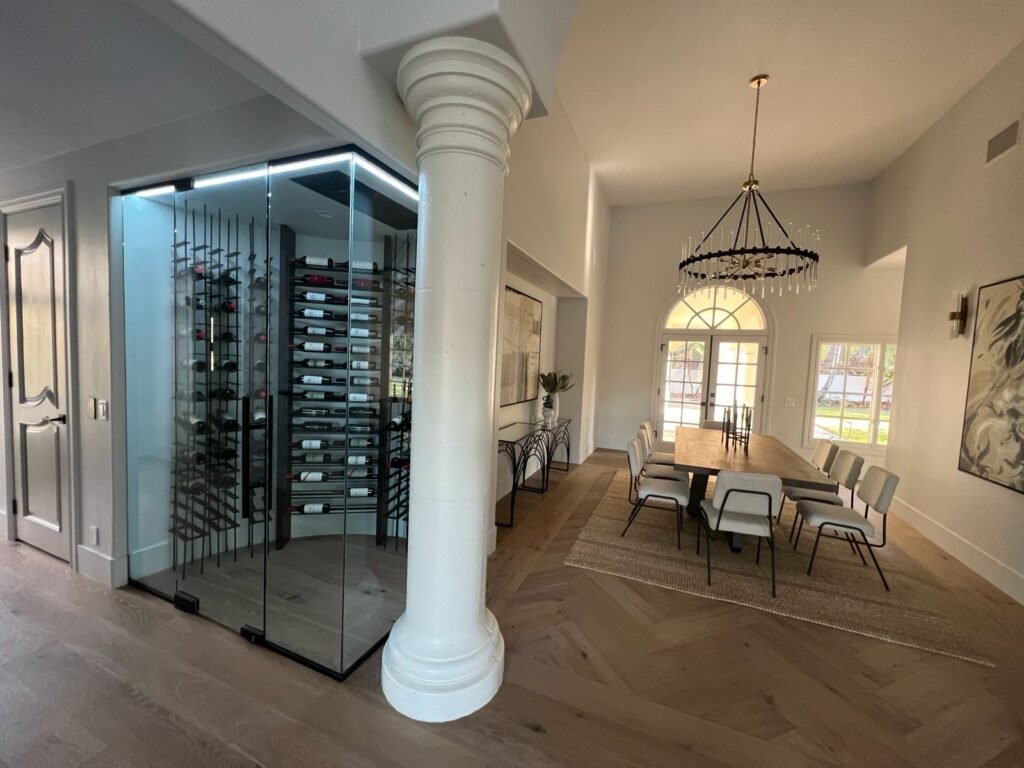 Glass-enclosed wine display next to a white column, with a dining table and chairs under a chandelier in a bright room with arched doorway — Construction Crafters project.