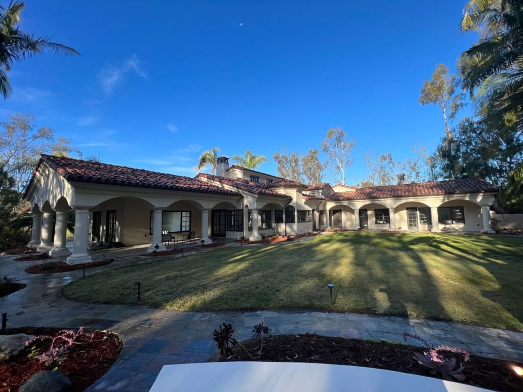 Rear view of a large Mediterranean-style home with a covered colonnaded patio, expansive green lawn, stone walkway, and mature landscaping under a clear blue sky