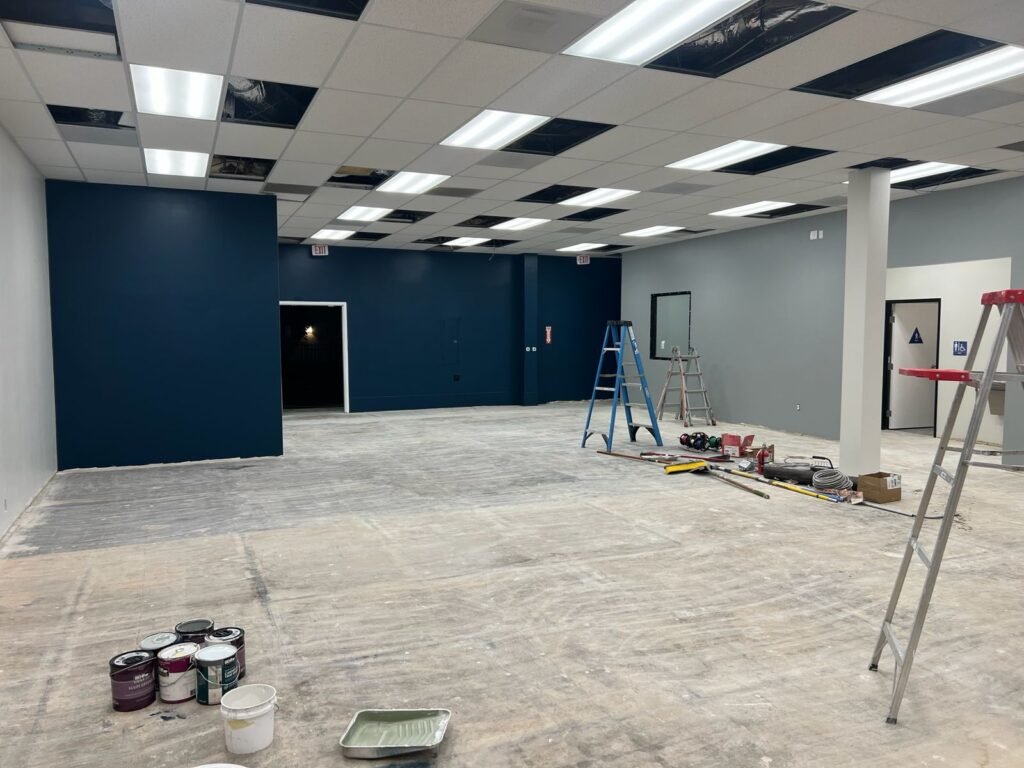Large commercial interior under renovation with a partially installed drop ceiling, ladders, paint cans, and tools scattered on the unfinished floor; walls painted navy blue and gray; exit door and restroom visible in background — Construction Crafters project.