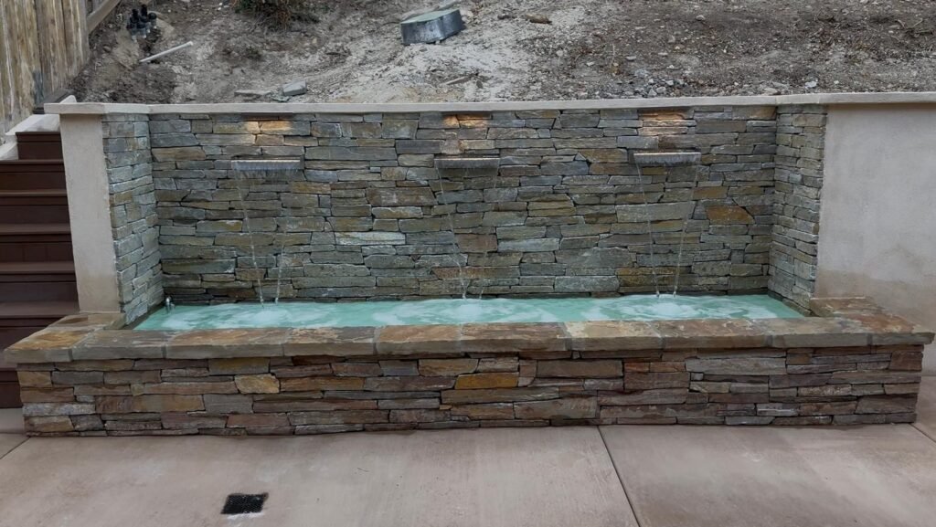 Stone water feature with three narrow spouts cascading into a long rectangular basin, built of stacked stone, with steps visible to the left and bare soil above — Construction Crafters project.