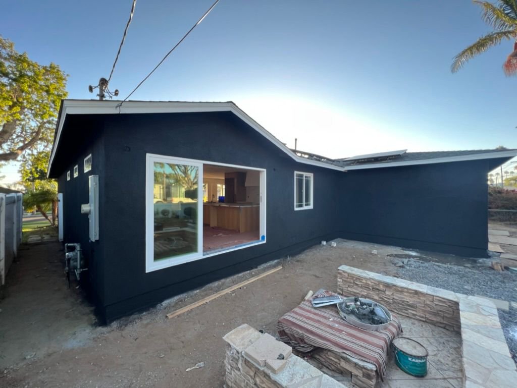Dark blue stucco home exterior mid-renovation with a large new window opening, stone low wall in the foreground, and yard prep materials.