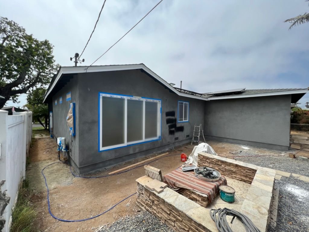 Gray stucco home exterior mid-renovation with masked windows, new stone low wall, and yard prep materials.