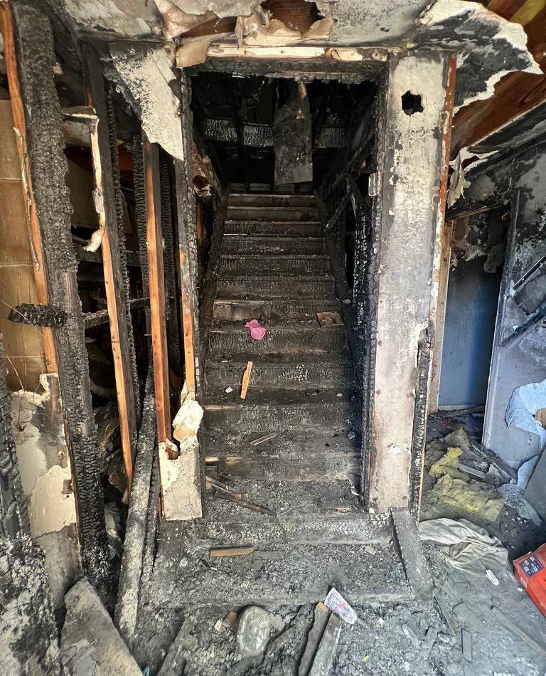 Interior stairwell and walls severely damaged by fire, showing charred wood framing, burnt drywall remnants, and debris-covered steps.