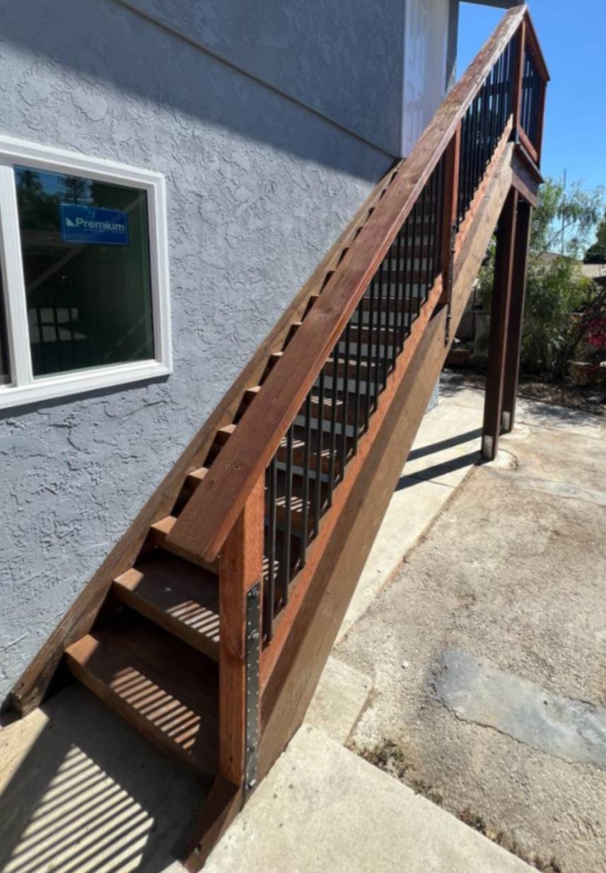 Exterior wooden staircase with black metal balusters leading to an upper-level door on a gray stucco building.