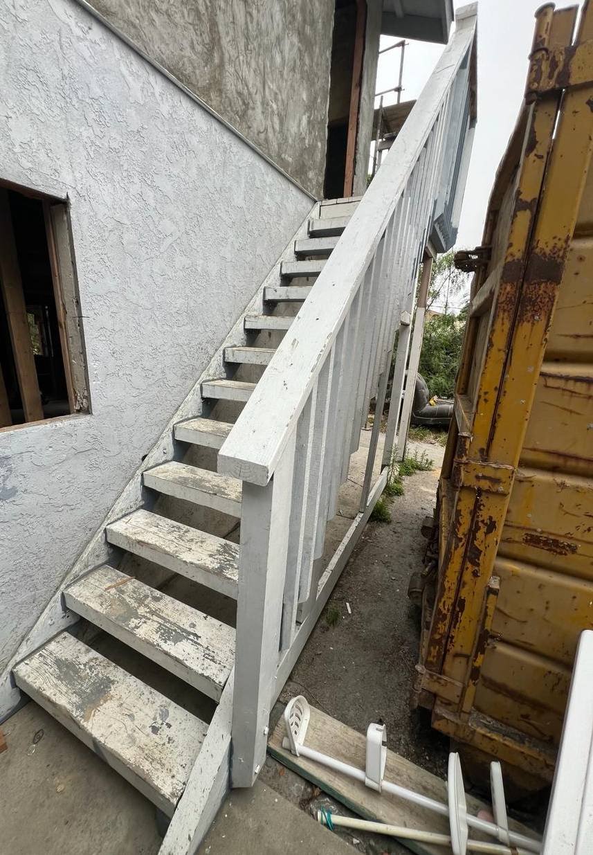 Worn exterior wooden staircase with peeling white paint attached to a stucco wall, showing signs of weathering and surrounded by renovation debris.