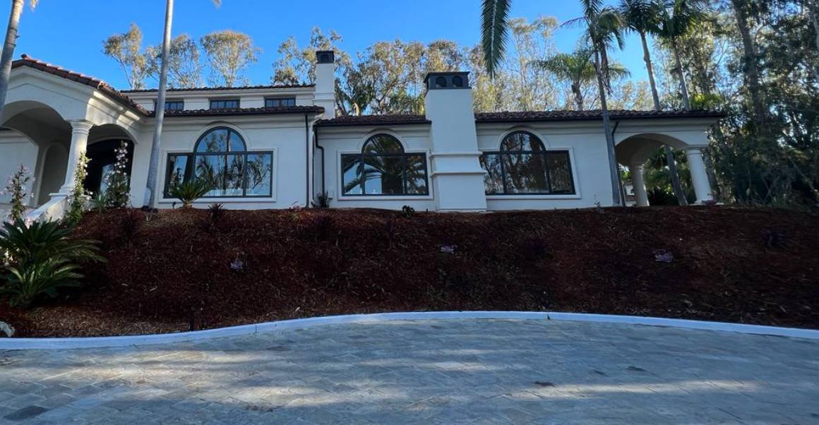 Mediterranean-style stucco home with refreshed landscaping, mulched hillside, and stamped concrete driveway under a clear sky.