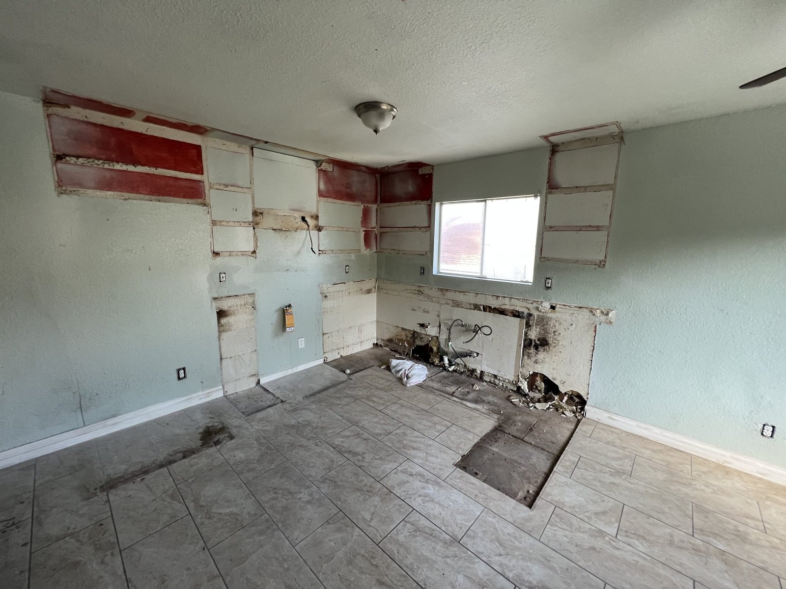 Kitchen area mid-demo with removed cabinets, exposed plumbing and wall cavities, and new tile floor partially patched.