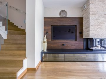 Modern living room corner with wooden stairs, wall-mounted TV on dark wood panel, low console, and a stone fireplace — Construction Crafters home remodeling