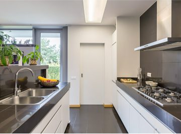 Narrow modern galley kitchen with white cabinets, dark countertops, stainless steel range hood, and plants by a window — Construction Crafters home remodeling