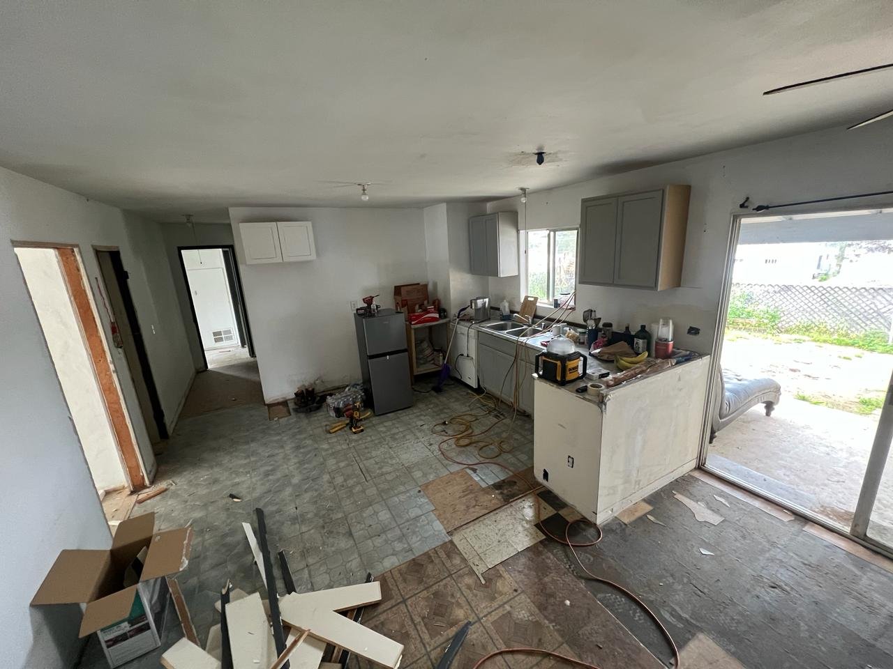 Open-concept kitchen/living area mid-renovation with torn-out flooring, demo debris, exposed subfloor, partially installed gray cabinets, and temporary appliances.