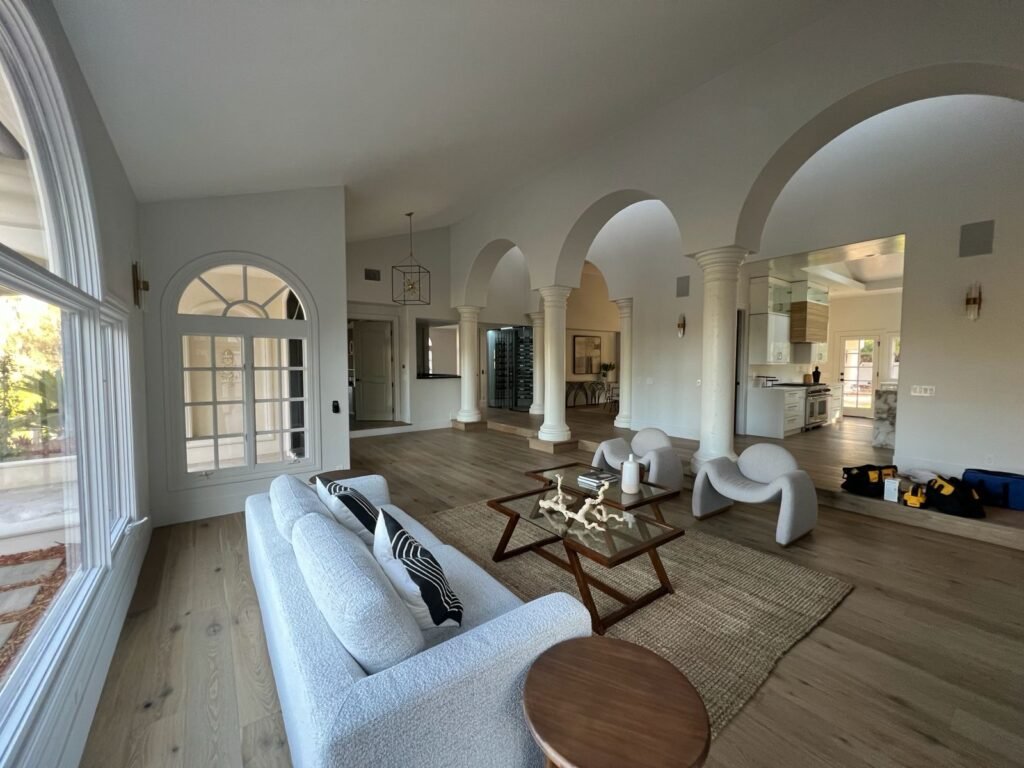 Spacious living room with white columns and arches, modern seating on a rug, large windows on the left, and a view into an adjacent kitchen — Construction Crafters project.