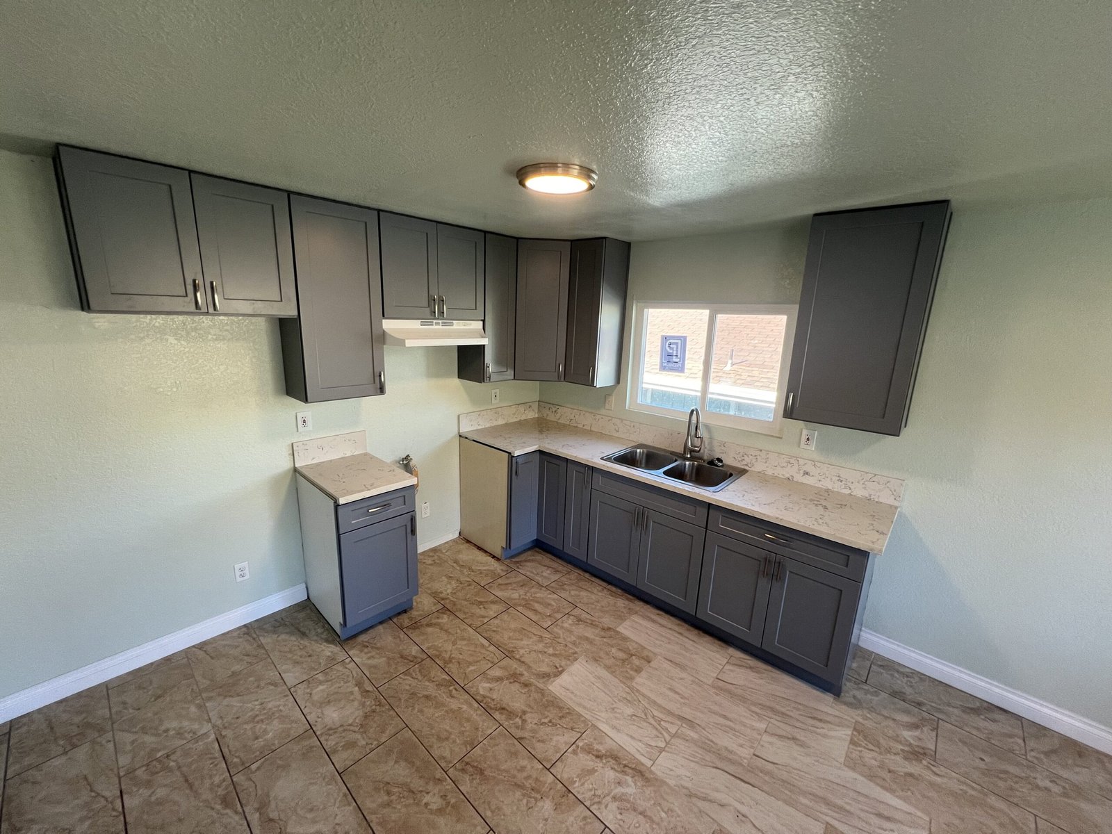 Updated kitchen with gray shaker cabinets, quartz countertops, tile flooring, and stainless-steel sink under a window.