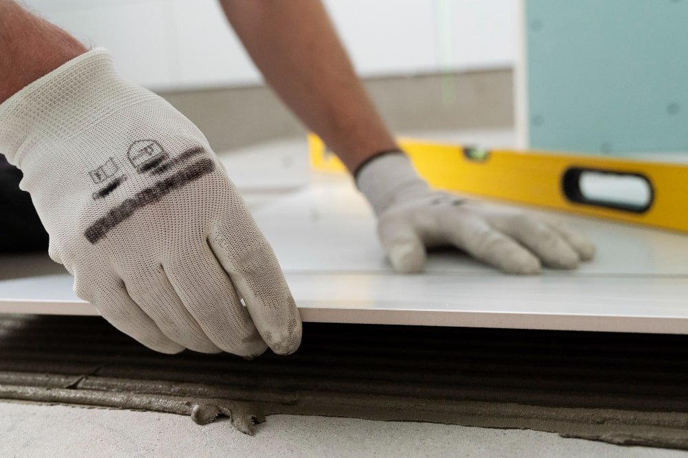Worker’s gloved hands pressing a tile onto gray mortar with a yellow level in the blurred background — Construction Crafters home remodeling