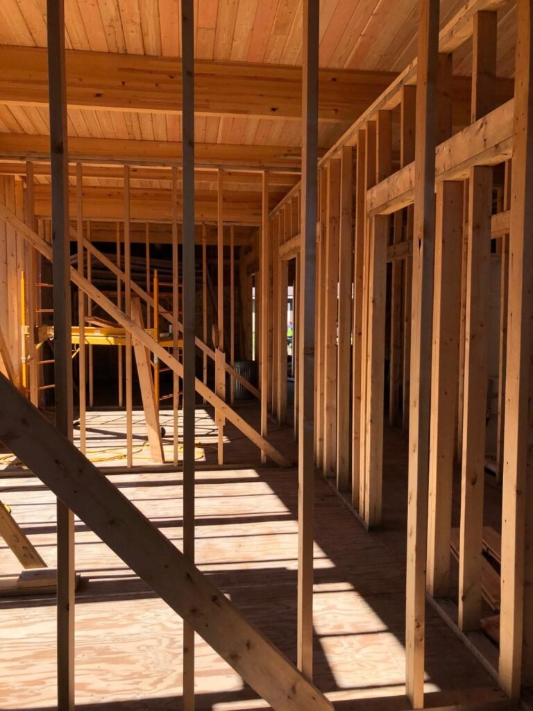 Two-story wooden house frame under construction surrounded by trees, with scaffolding on the left and building materials in the foreground. Construction Crafters
